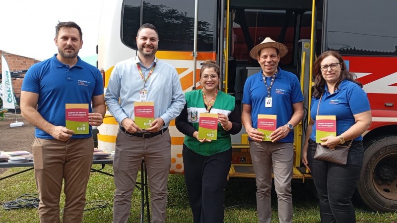 Em frente ao novo ônibus do Procon estão cinco pessoas segurando livros do Código de Defesa do Consumidor. As pessoas são: o prefeito de Três de Maio, Marcos Corso, de camiseta azul escura; o diretor-executivo do PROCON RS, Rainer Grigolo, de camisa azul clara; a Chefe De Divisão de Atendimento e Fiscalização da entidade, Barbara Ramos, vestindo um colete do Procon; o vice-prefeito de Três de Maio, Josias Correa, de azul escuro e chapéu panamá; e uma servidora do município de Três de Maio, de camiseta azul escura.
