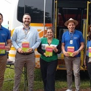 Em frente ao novo ônibus do Procon estão cinco pessoas segurando livros do Código de Defesa do Consumidor. As pessoas são: o prefeito de Três de Maio, Marcos Corso, de camiseta azul escura; o diretor-executivo do PROCON RS, Rainer Grigolo, de camisa azul clara; a Chefe De Divisão de Atendimento e Fiscalização da entidade, Barbara Ramos, vestindo um colete do Procon; o vice-prefeito de Três de Maio, Josias Correa, de azul escuro e chapéu panamá; e uma servidora do município de Três de Maio, de camiseta azul escura.